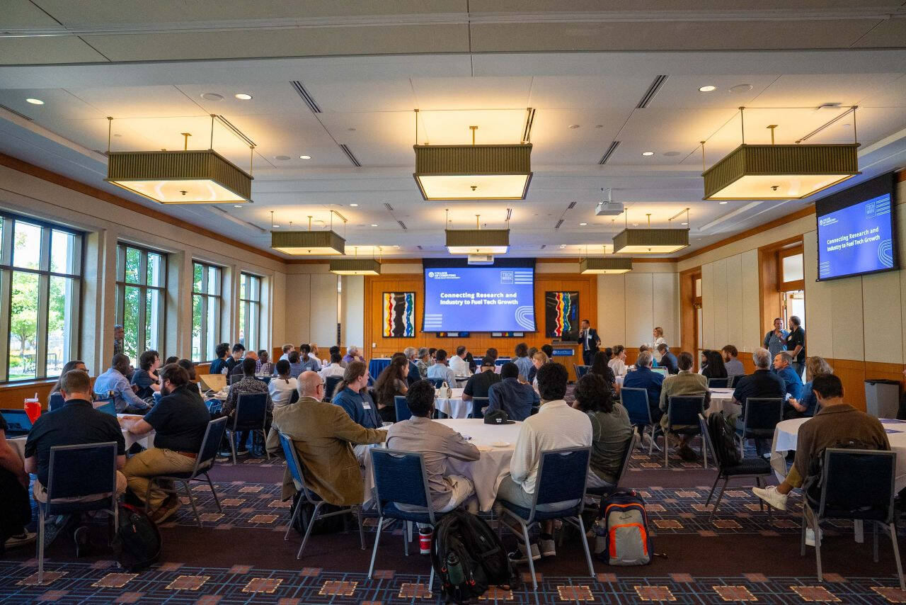 A large audience sits at round tables inside the DeVos Center listening to a Tech Week Grand Rapids session titled “Connecting Research and Industry to Fuel Tech Growth.” Presentation slides are displayed on large screens at the front of the room.